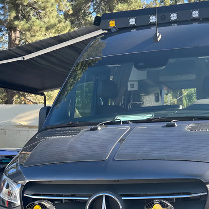 Front view of a Mercedes-Benz Sprinter camper van with the Vantrix manual side awning extended at a wooded campsite.